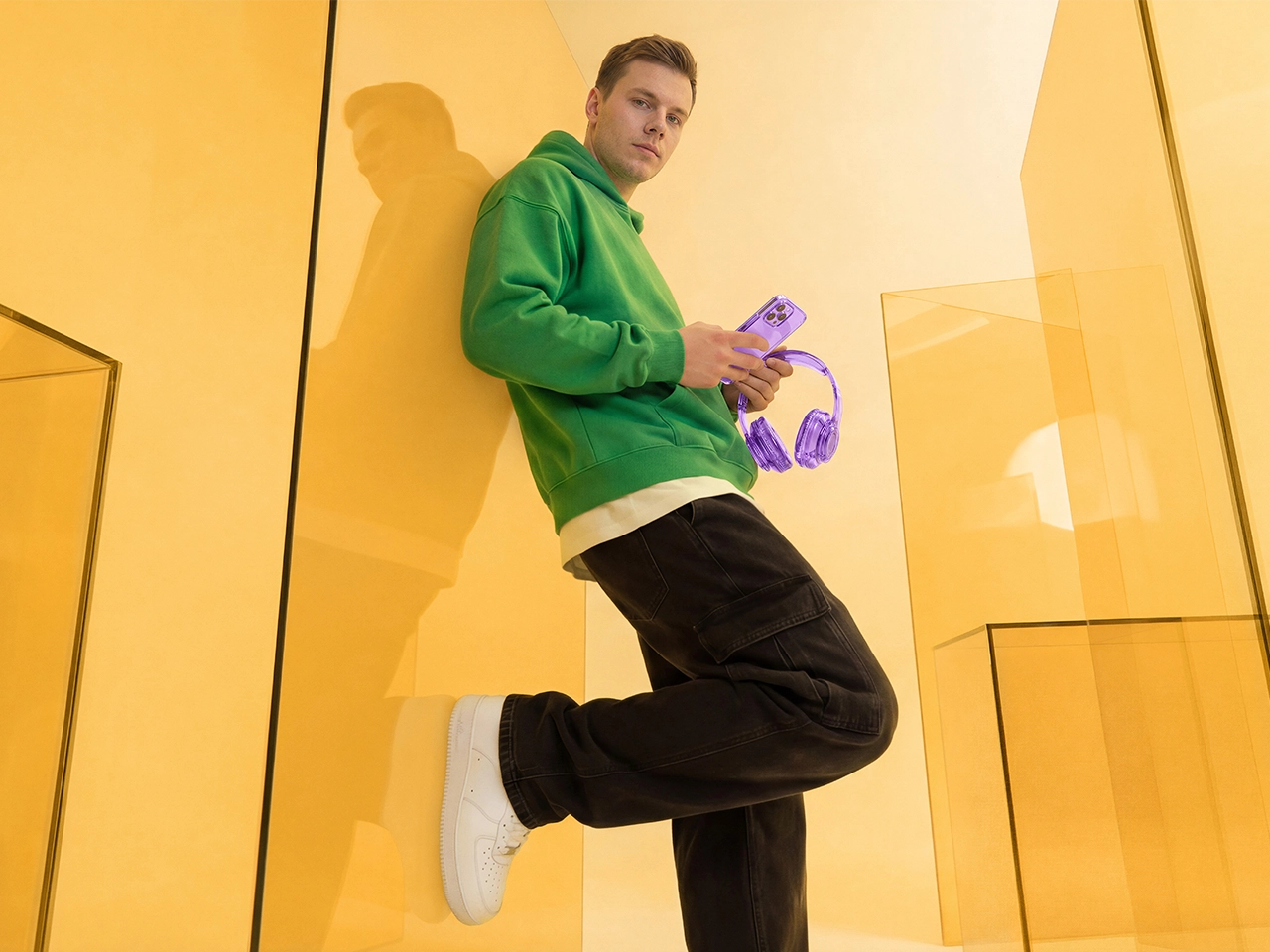 A man in a green hoodie and black cargo pants leans against a yellow translucent wall, holding a purple smartphone and purple headphones.