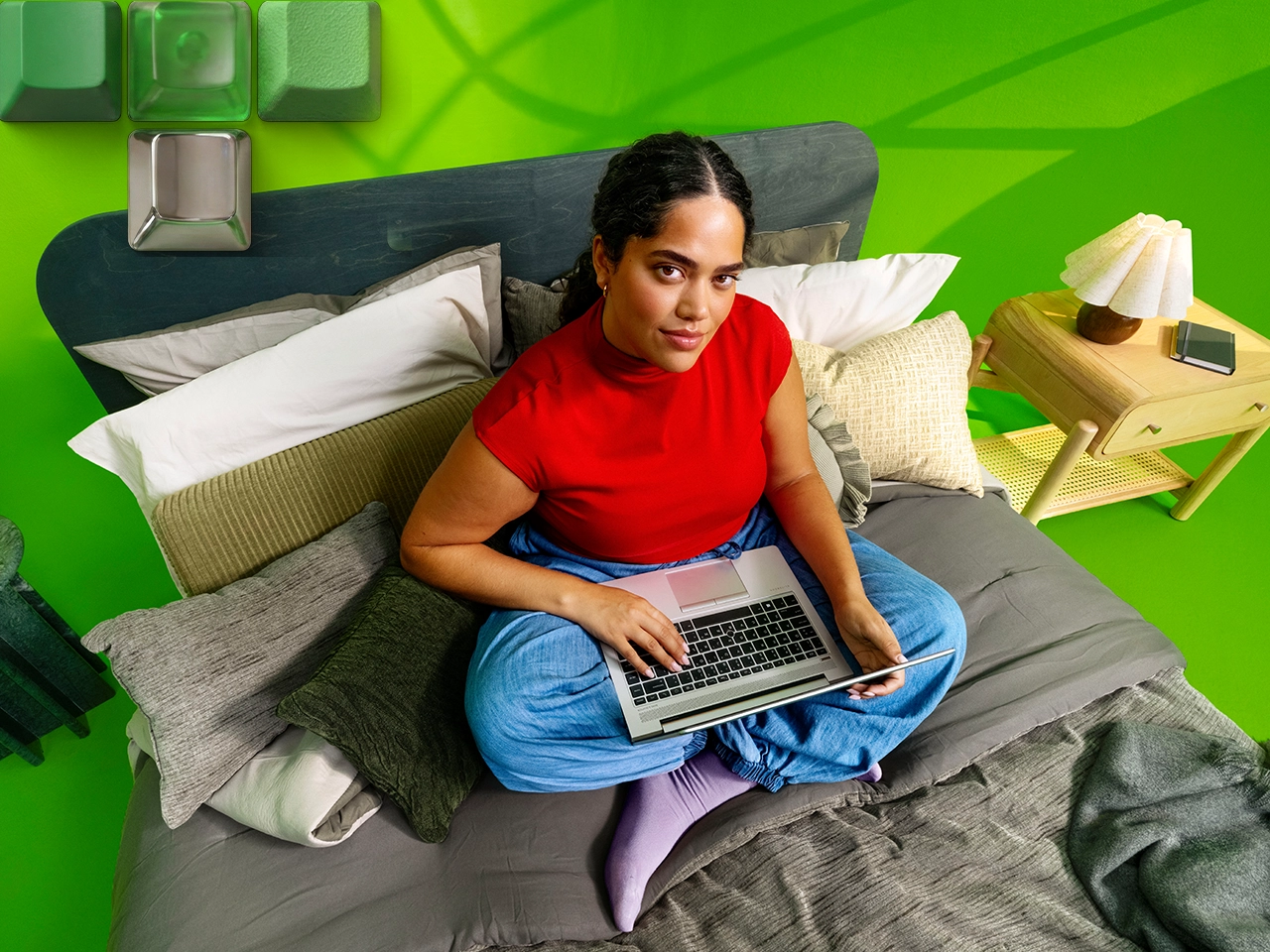 Woman sitting cross-legged on bed using laptop, bright green room, large computer keys.