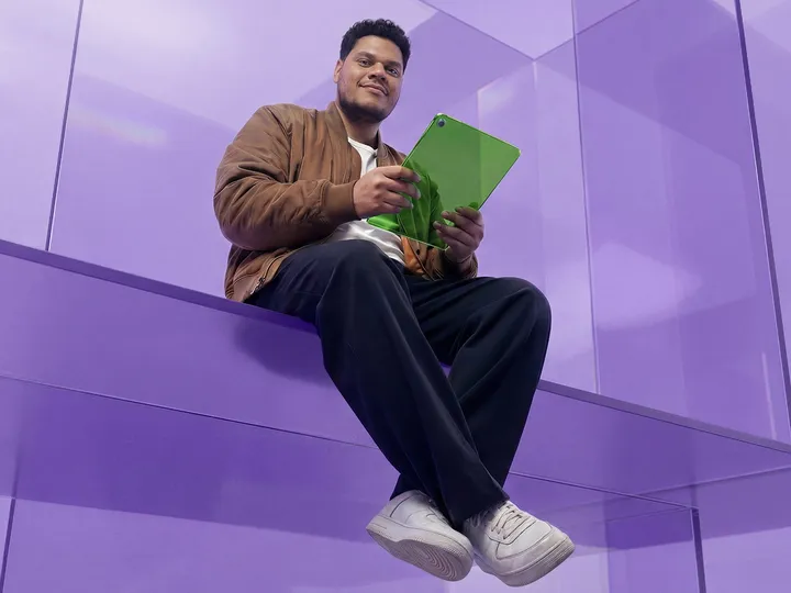 A low-angle shot of a smiling man in a brown jacket sitting on a purple ledge while holding a vibrant green tablet.