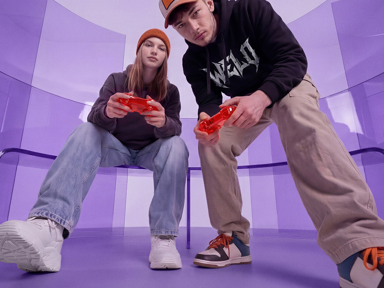 Two people sitting on a bench, holding red game controllers, focusing on a video game in a modern, purple-toned room.