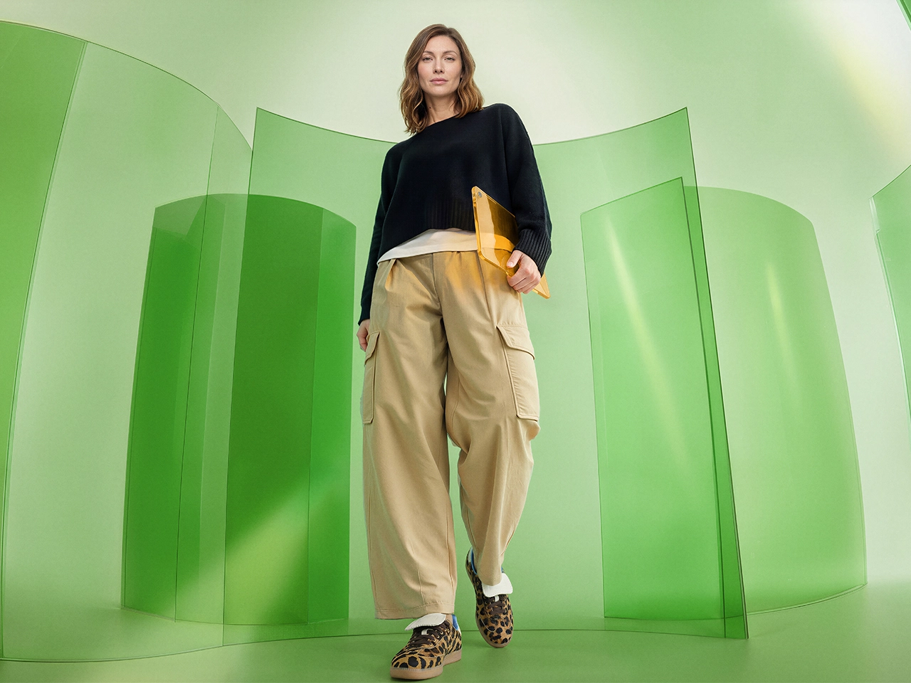 Low-angle woman in black sweater, tan cargos walks through green room with curved panels, carrying yellow laptop