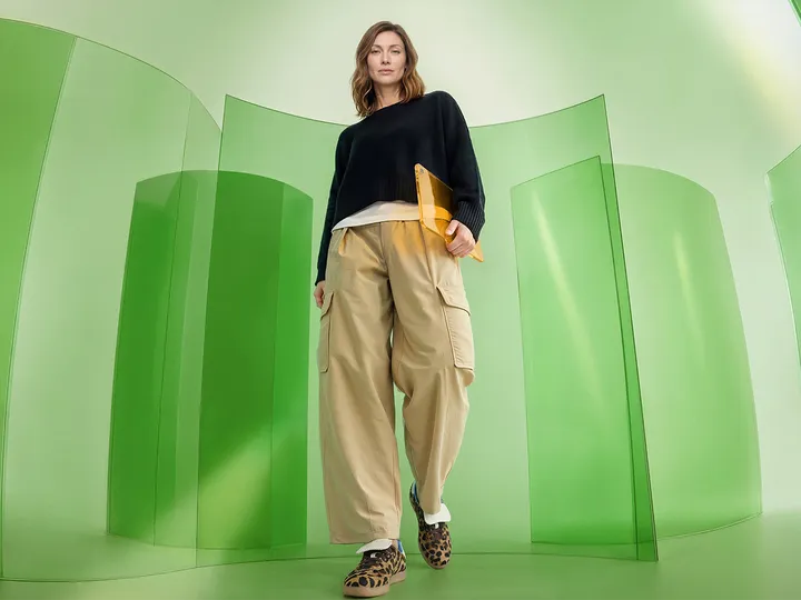 Low-angle woman in black sweater, tan cargos walks through green room with curved panels, carrying yellow laptop.