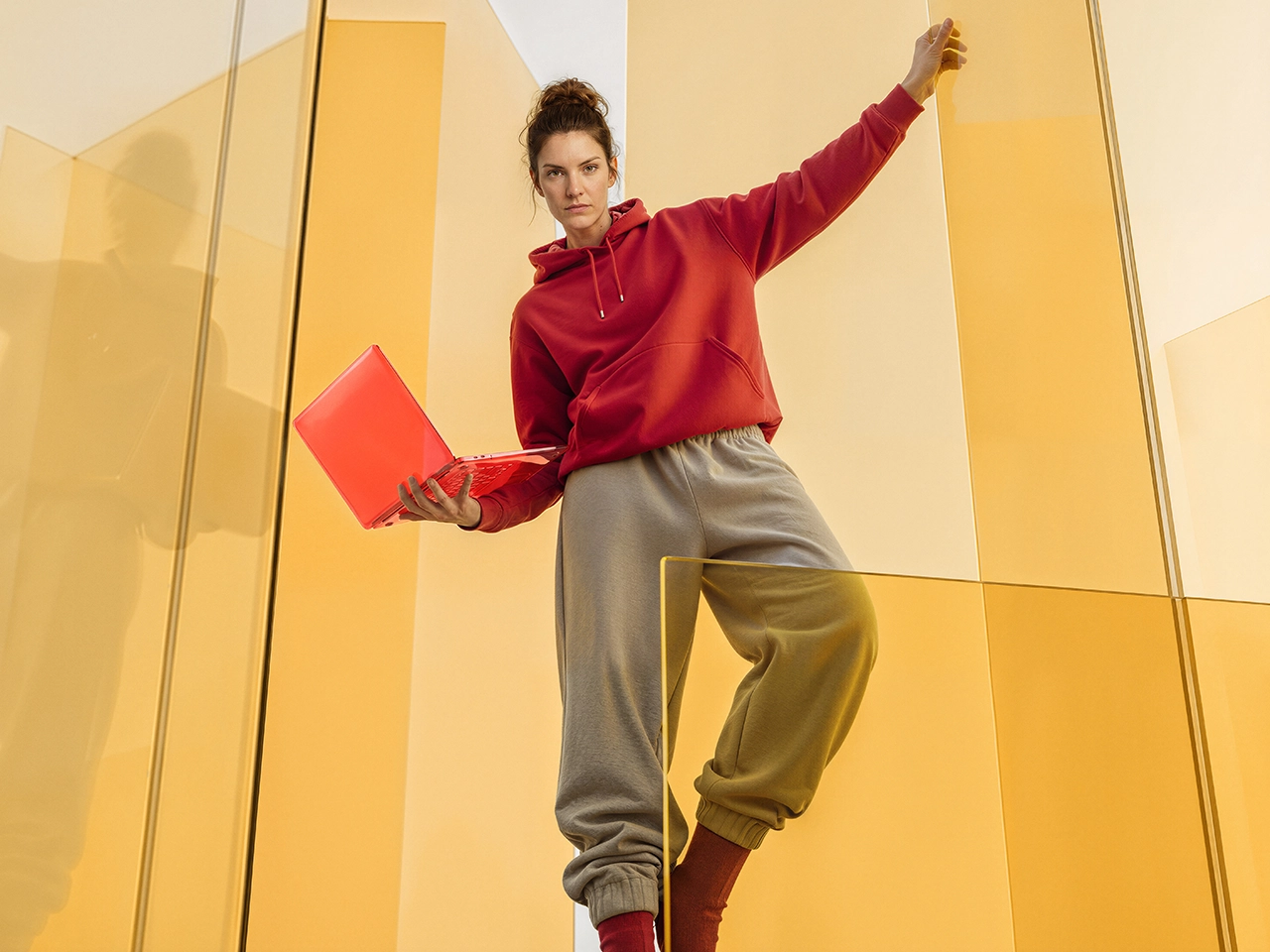 A woman in a red hoodie and tan pants holding a red laptop in a yellow room.