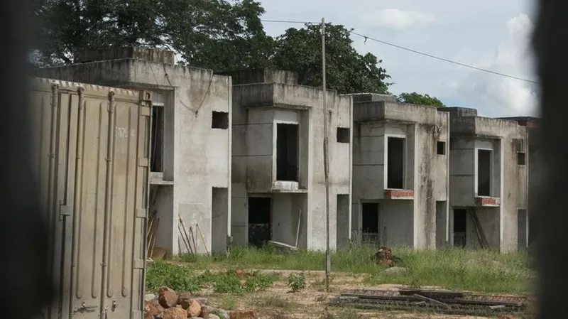 Asas inacabadas em estado de abandono, com paredes de concreto expostas, sem portas ou janelas instaladas. O terreno ao redor está coberto por vegetação rasteira e entulhos, com um contêiner metálico à esquerda. O céu está parcialmente nublado.