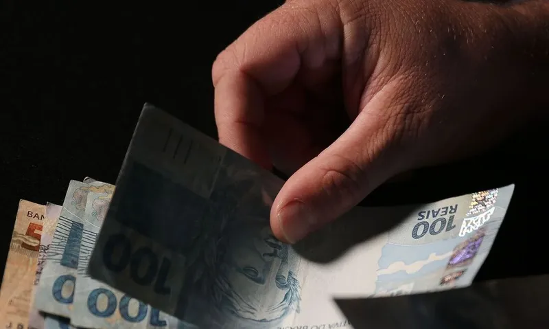 AltText: Mão masculina segurando notas de R$ 200,00, R$ 100,00, R$ 50,00 e R$ 20,00