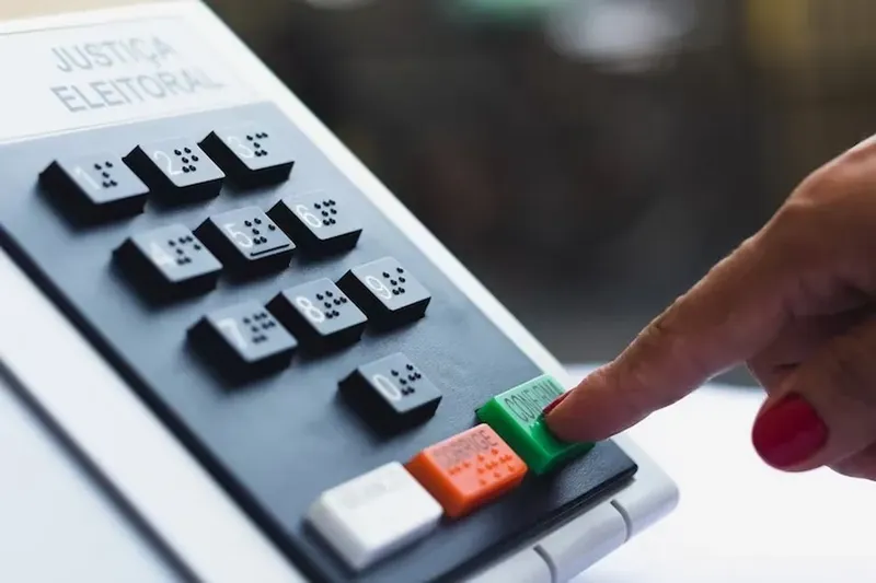 Uma urna eletrônica cinza com uma tela na parte superior e um teclado numérico abaixo com uma mão tocando a urna eletrônica.