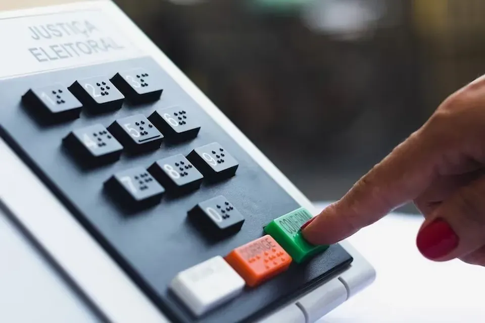 Uma urna eletrônica cinza com uma tela na parte superior e um teclado numérico abaixo com uma mão tocando a urna eletrônica.