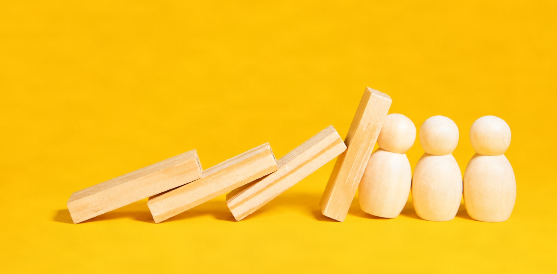 Wooden blocks tipping over like dominos against three wooden figurines on a bright yellow background.
