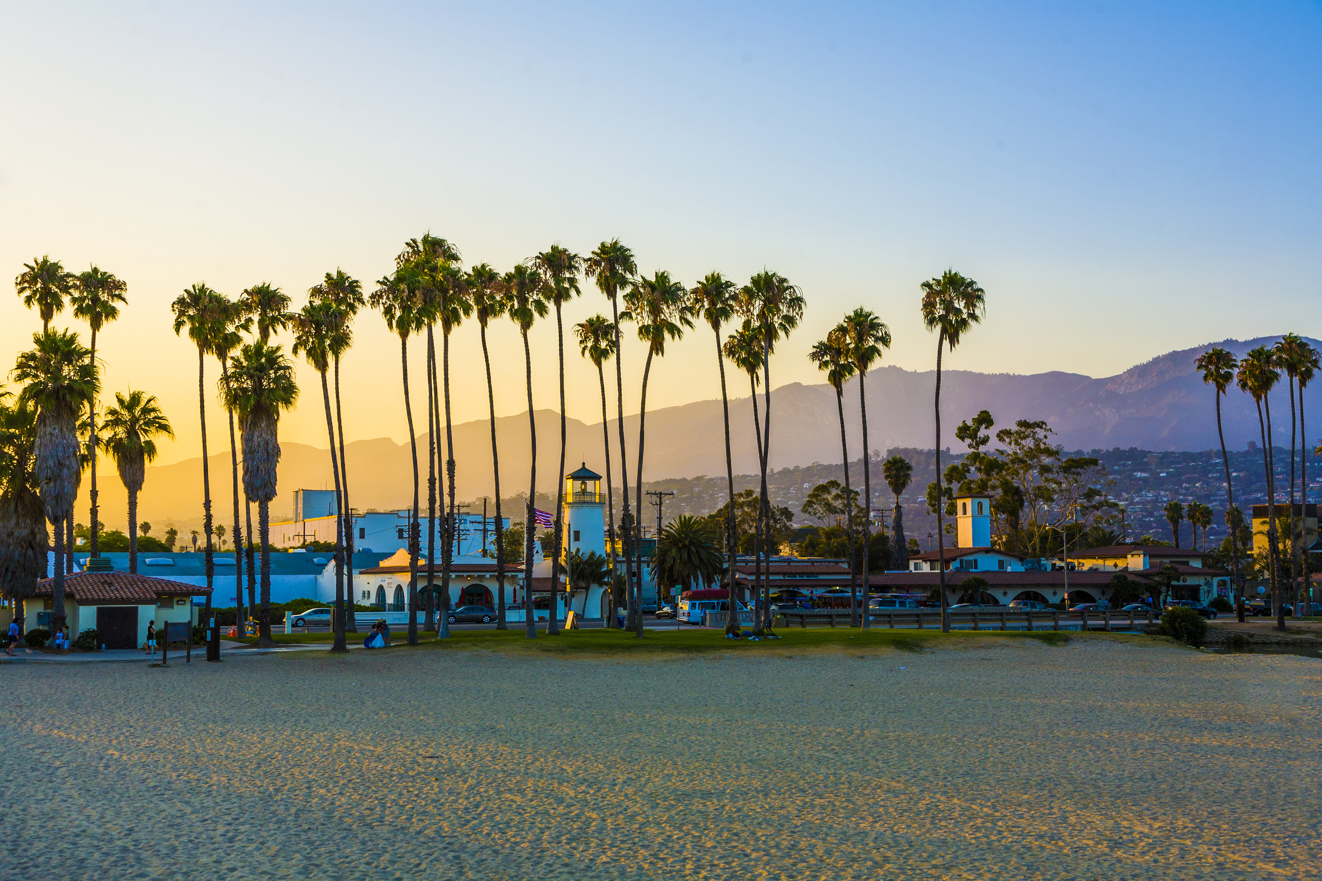 Sunset over a beach with tall palm trees, a lighthouse, and buildings, framed by mountains in the background.