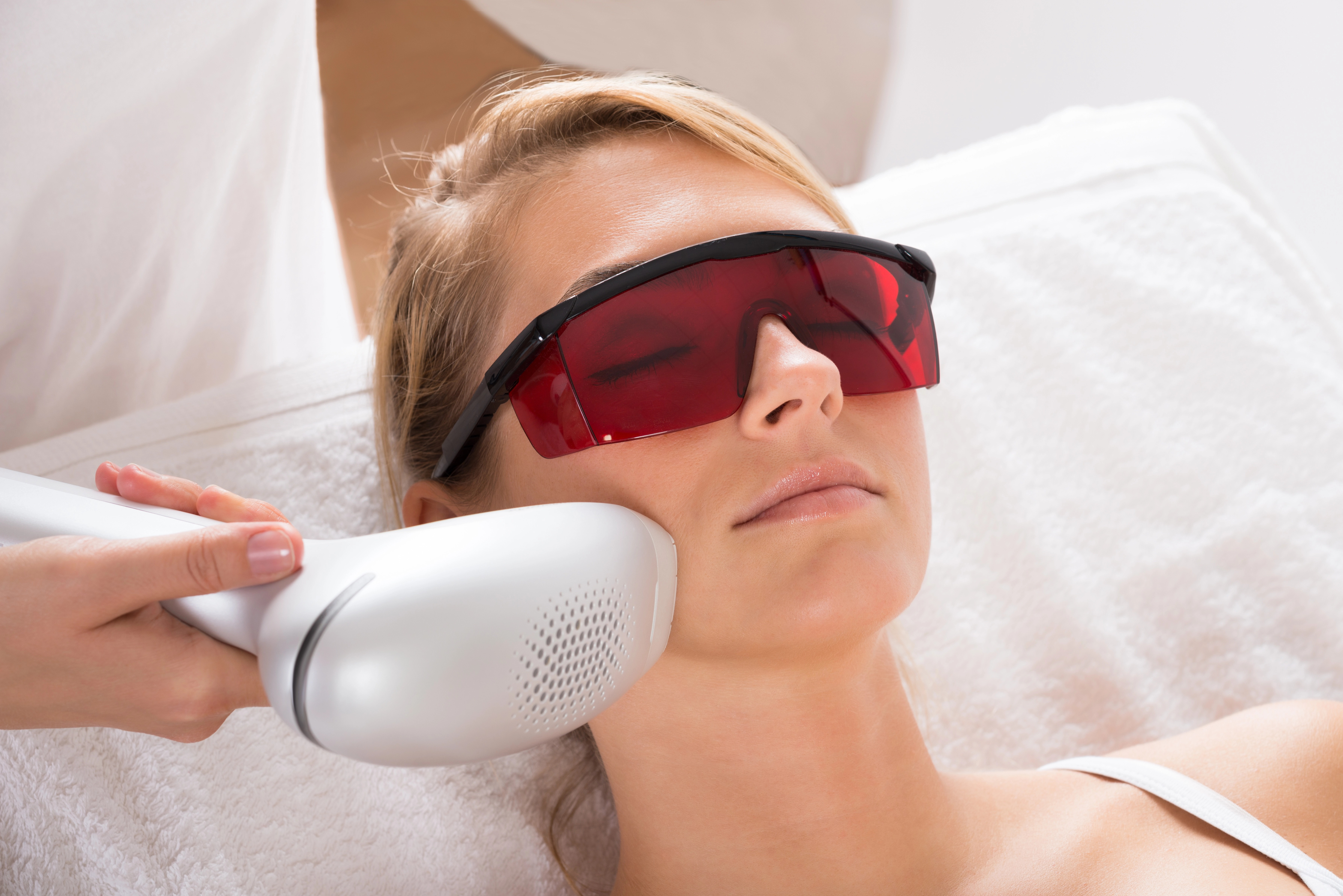 Person receiving a facial laser treatment while wearing protective red goggles, technician holding a laser device to their cheek.