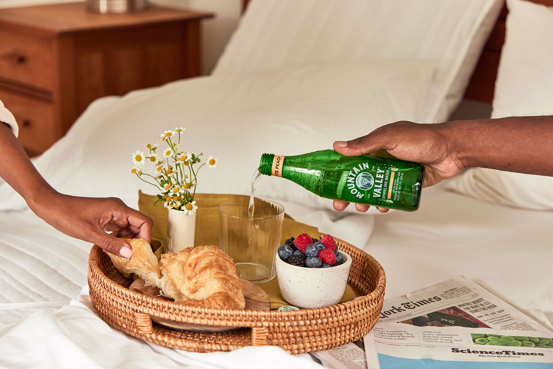 A breakfast tray with a croissant, berries, a small vase of flowers, and a person pouring sparkling water into a glass on a bed.