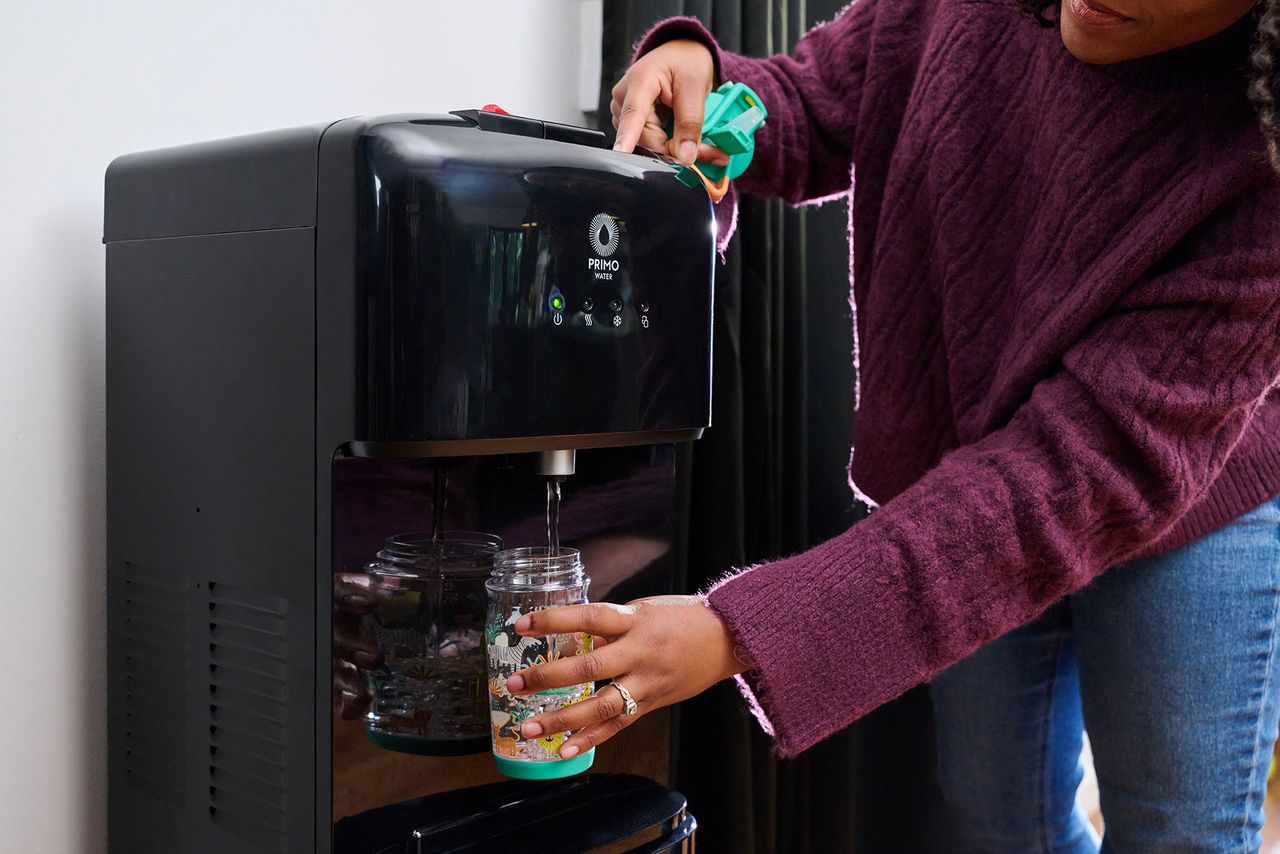 A person in a purple sweater fills a mason jar with water from a black Primo water dispenser.