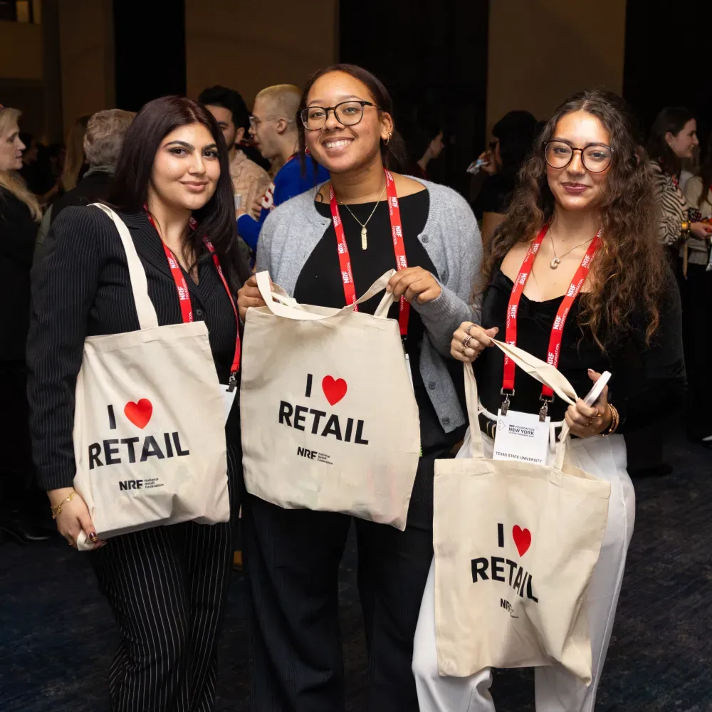 Three women smiling and holding tote bags reading "I ♥ RETAIL" at a networking event, wearing red lanyards with ID badges.