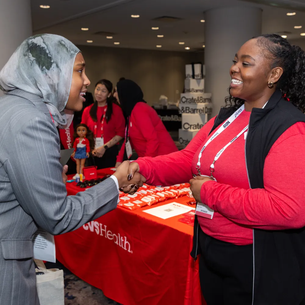 Two women shaking hands and smiling at a CVS Health event booth, with others in red attire in the background.