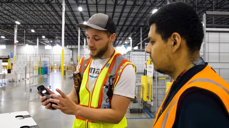Two workers in an Amazon warehouse are seen interacting. One worker is holding a handheld device and appears to be explaining or demonstrating something to the other worker.