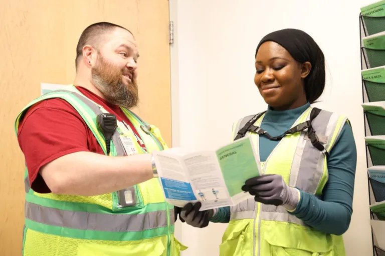 Two individuals wearing high-visibility safety vests are standing indoors. One person is holding a booklet and pointing at it, while the other person is looking at the booklet and holding it with gloved hands.