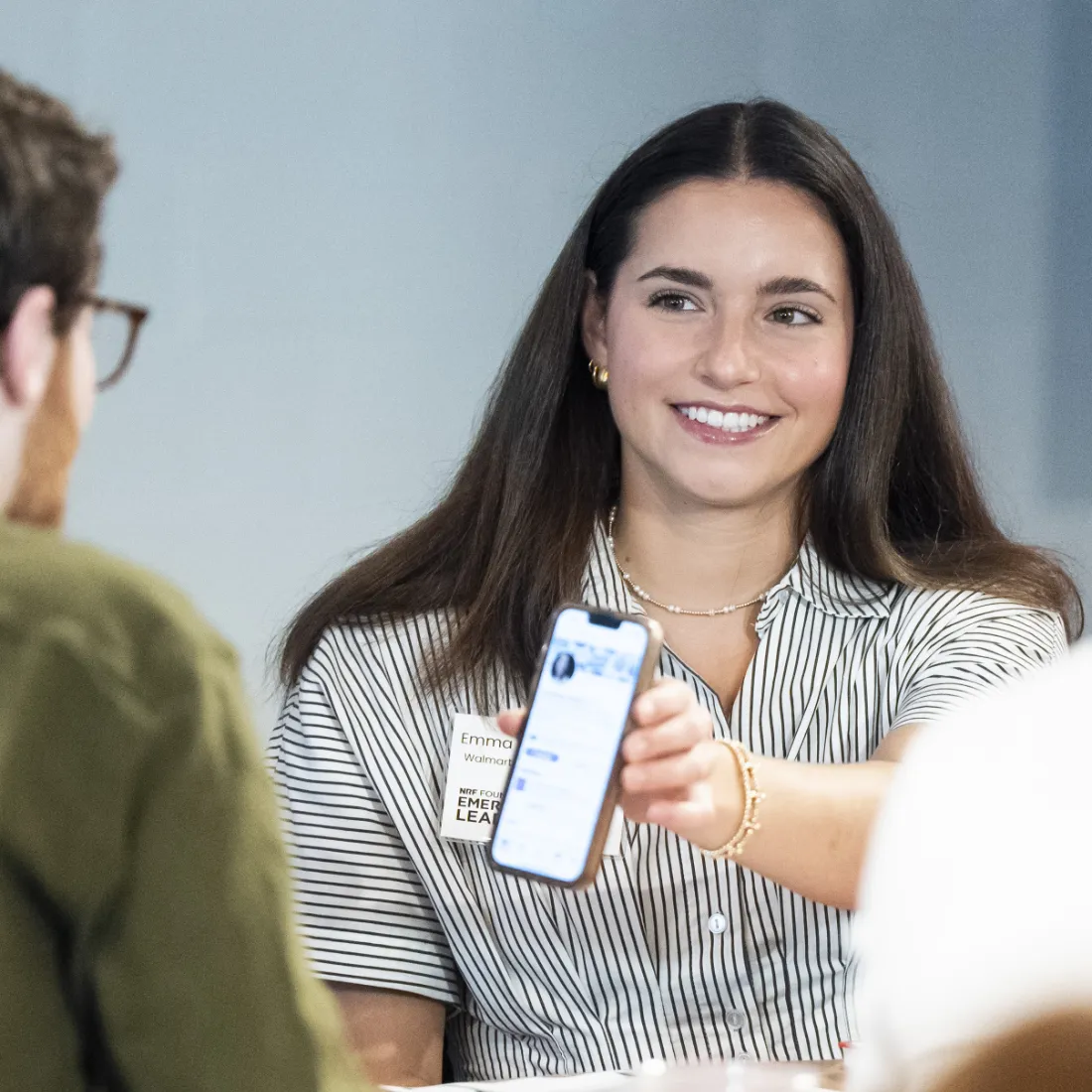 A young, smiling woman shows a young man the content of her phone's screen while seated at conference table.