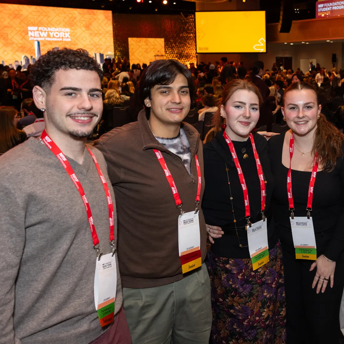 Four NRF NXT attendees smiling, wearing red lanyards with badges, pose together in a crowded conference room with a yellow screen in the background.