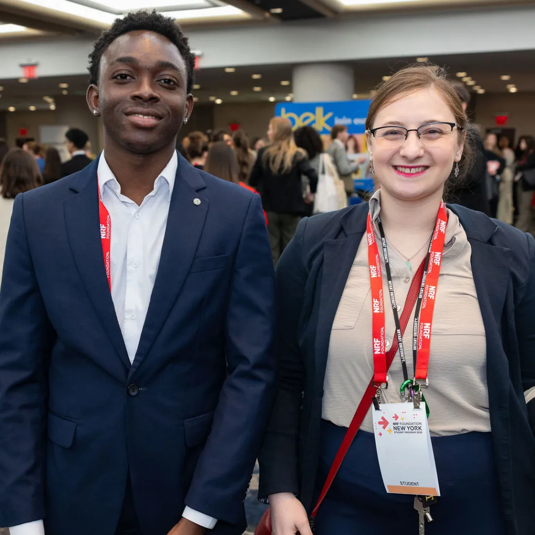 A man and woman, both wearing business attire and red lanyards, stand smiling at NRF NXT 2025 with people mingling in the background.