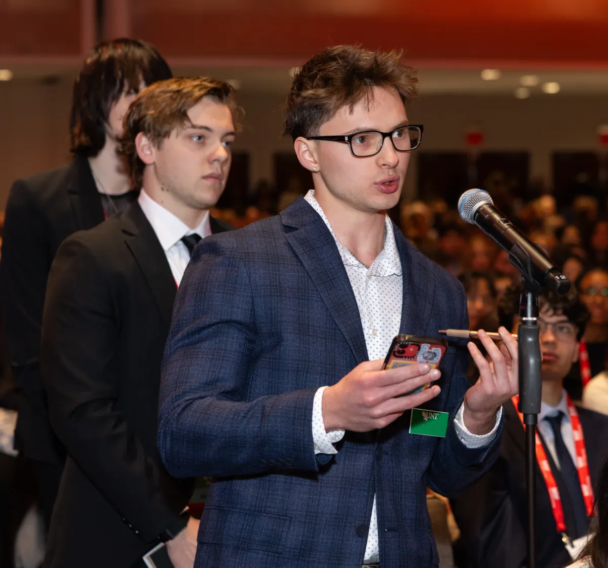 A young man wearing glasses speaks into a microphone at a NRF NXT, holding a phone and pen, with a crowd and other attendees in the background.
