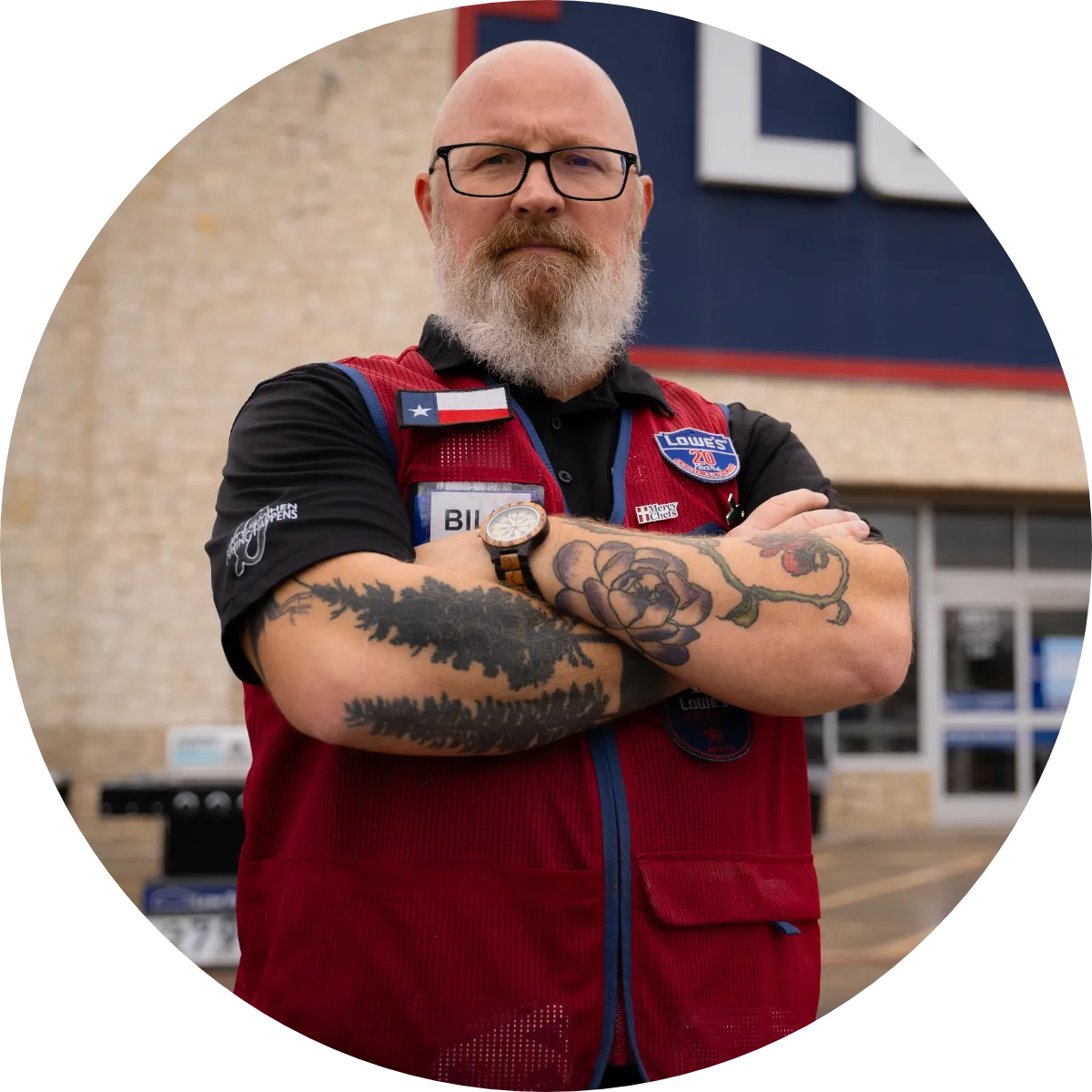 A representative from Lowe's poses with his tattooed arms crossed in front of the store wearing his Lowe's vest and nametag.