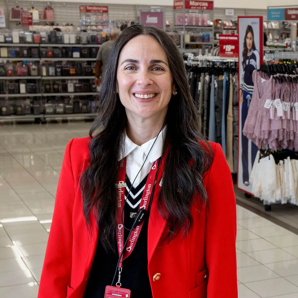 Elivette Sanabria wears a red blazer and lanyard and stands in a Burlington store with racks of clothes and shelves in the background.