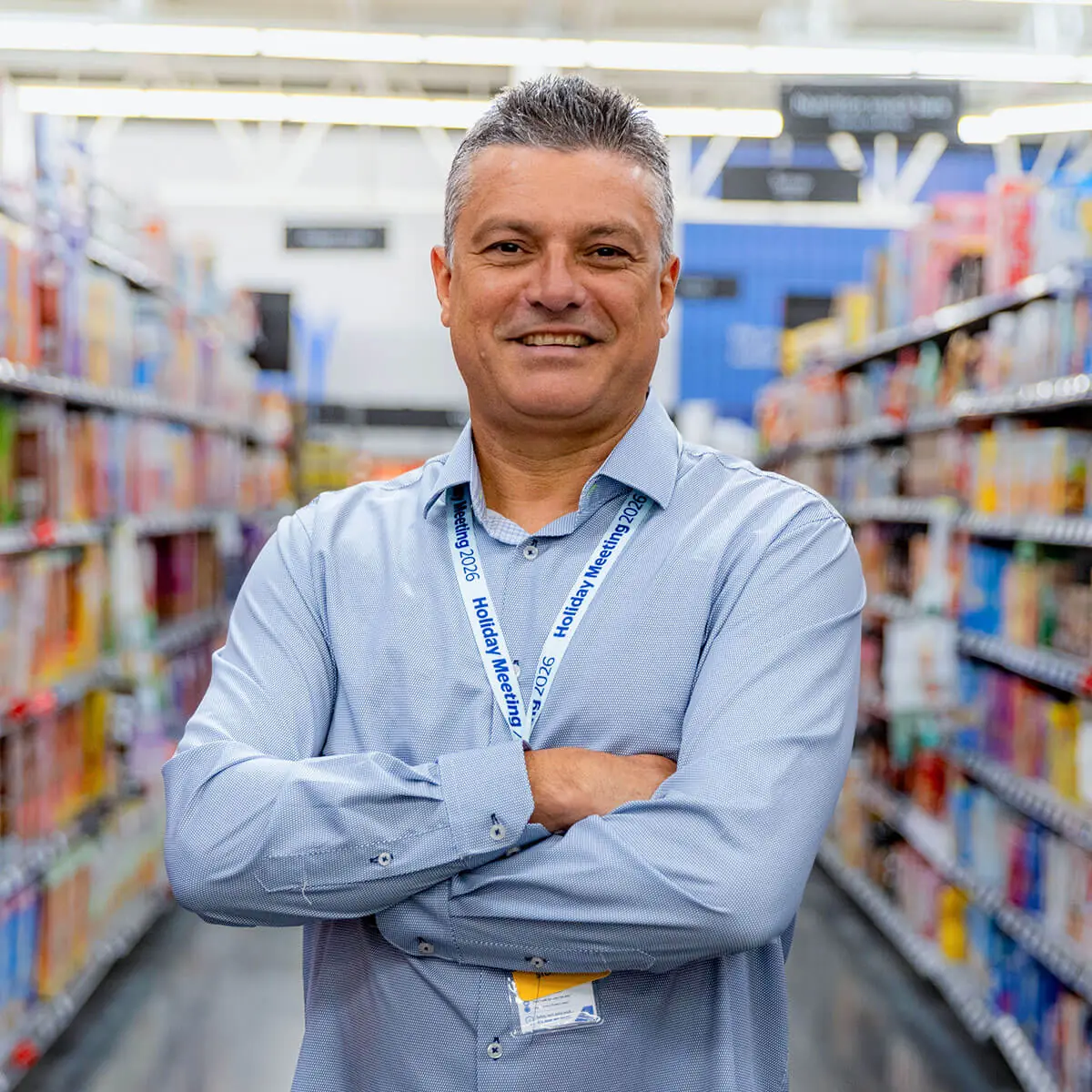 Juan Pinillos wearing a Walmart manager's blue shirt and a lanyard, standing with crossed arms in the aisle of a store filled with colorful products.