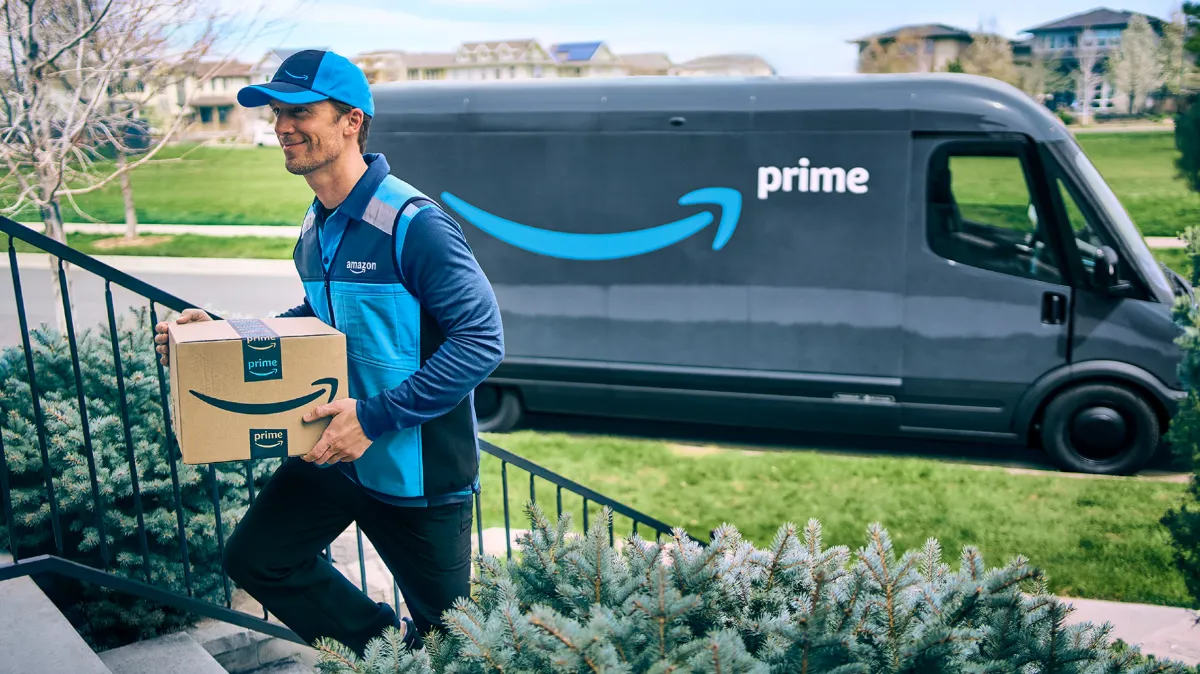 An Amazon truck driver is walking an Amazon package up a cement staircase. Behind him, parked along the curb, is a box truck with the Amazon smile arrow and the word Prime.
