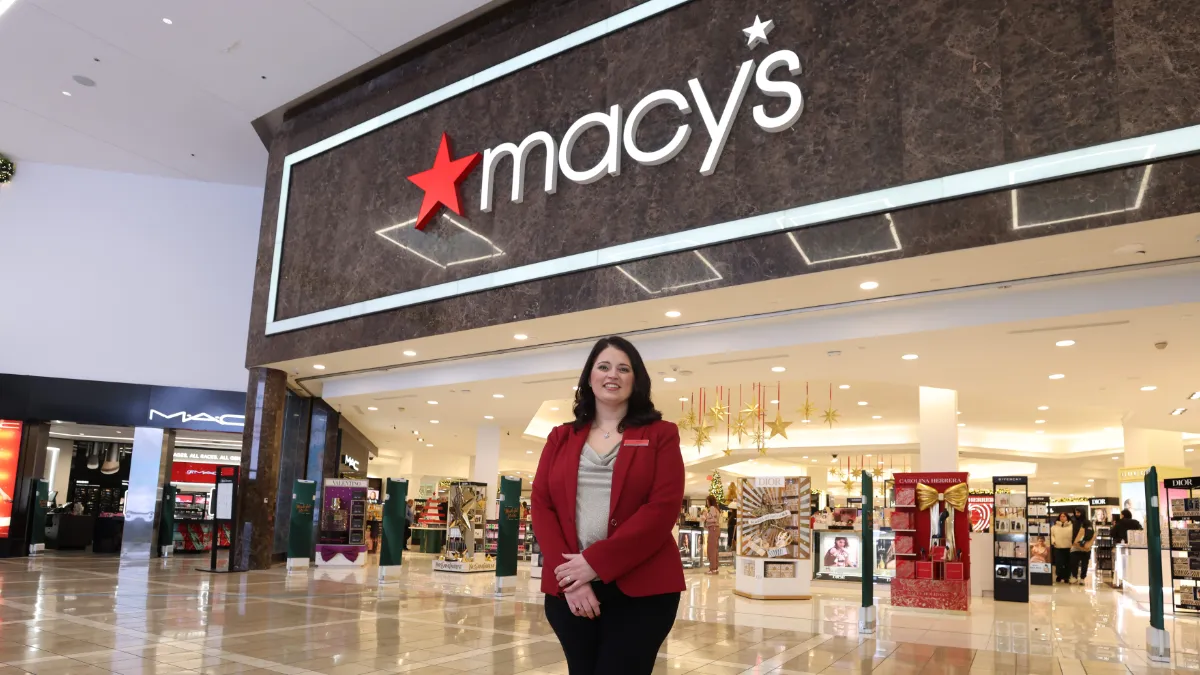 A Macy's Store Leader poses in front of the Macy's entrance, from within the mall. She's wearing a red blazer, and the store front behind is lit up and decorated with gold stars and gold ribbons