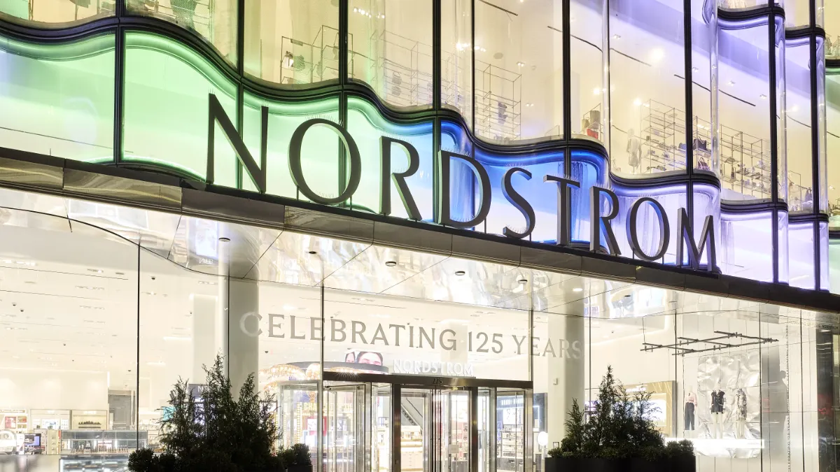 A photo of a Nordstrom store taken at night. The shelves and racks of the store are fully visible from the street under the department stores' bright lights. Above the store's revolving doors, a giant sign reading Nordstrom, glows in soft hues of green, blue, and purple.