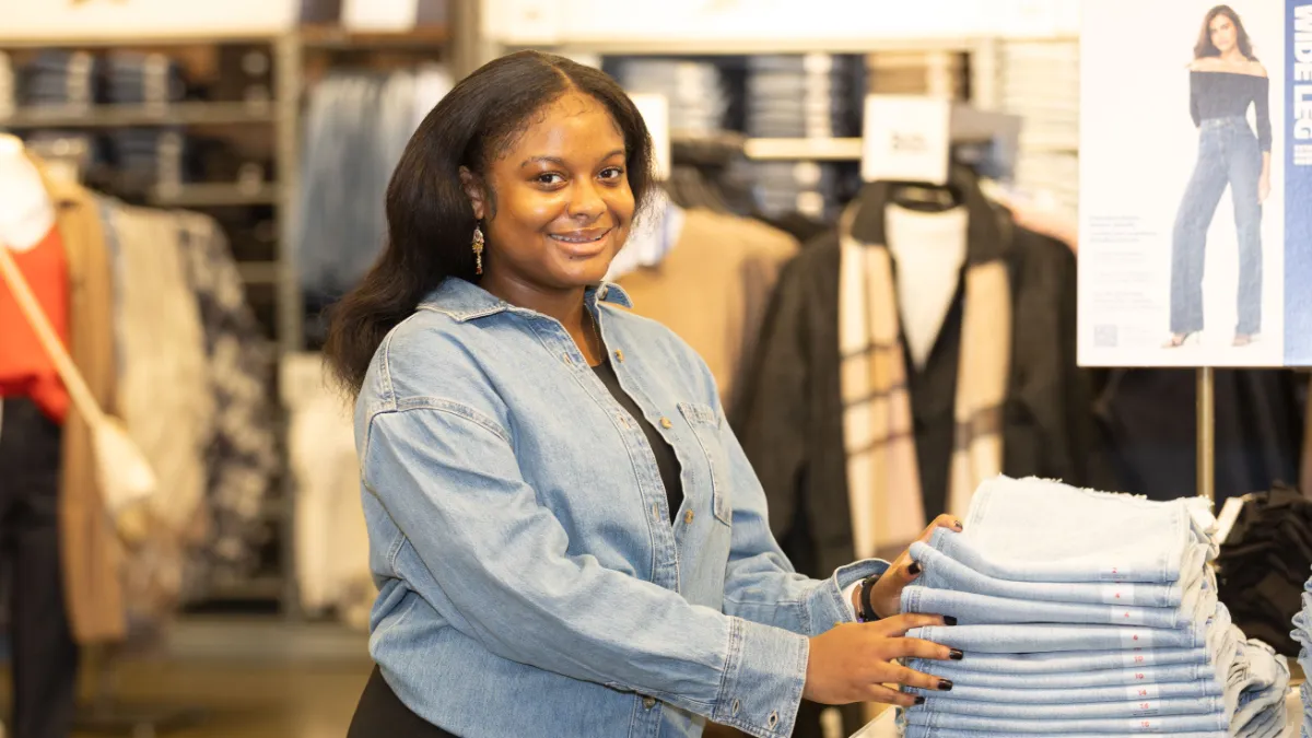 A woman in a light blue denim shirt smiles at the camera while managing a stack of Old Navy jeans.