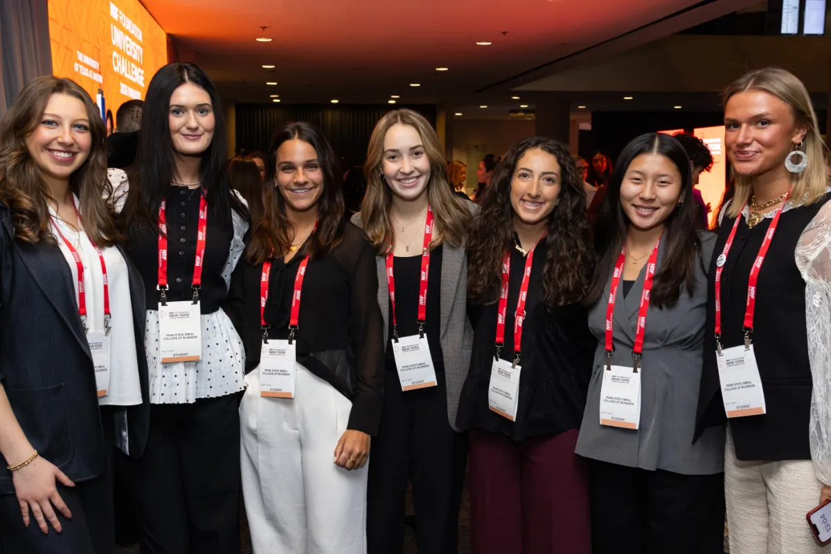 A group of seven women pose together at NRF NXT, wearing business attire and red lanyards. The background features an orange banner.