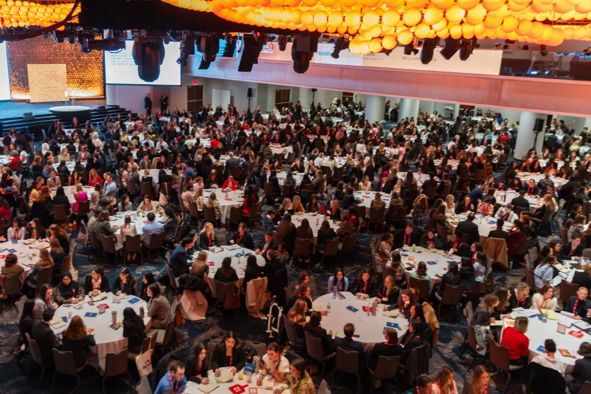 NRF NXT conference with numerous attendees seated at round tables, beneath a ceiling of decorative orange lights.