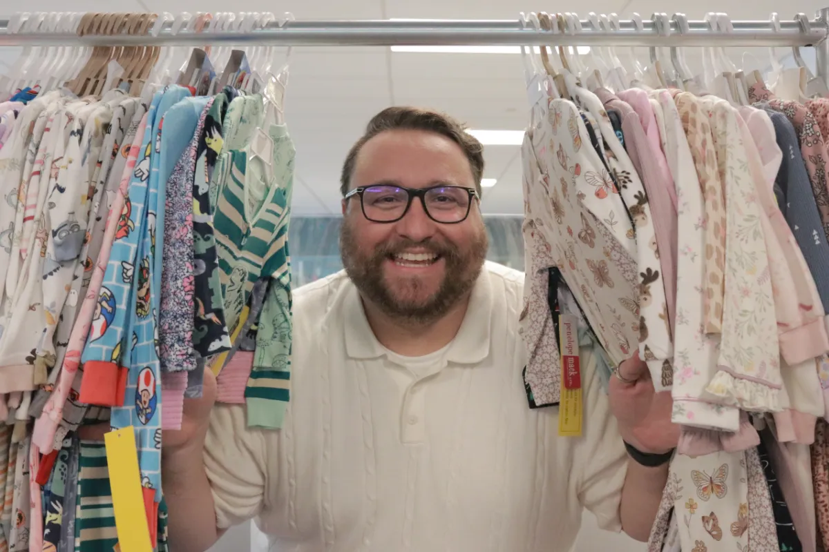 A man in fashionable glasses pushes his body through a rack of children's clothes with a big smile while in an office environment.