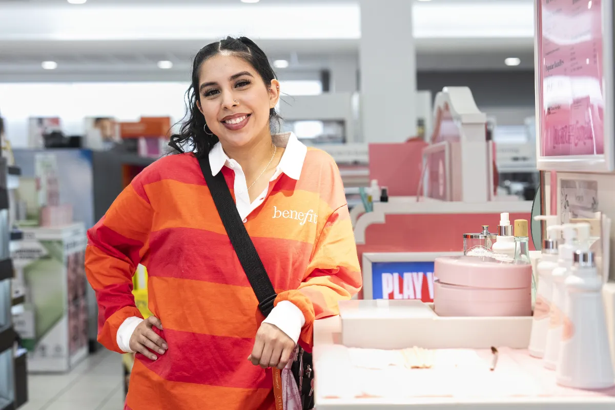 A woman with dark hair, wearing a red and orange rugby style shirt that says Benefit on it, leans on a store display of a bathroom sink environment on a department store showfloor.
