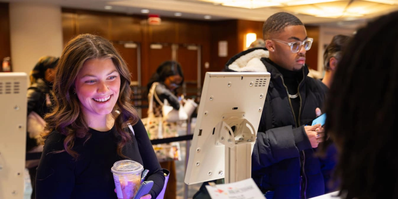 People at the NRF NXT registration counter, a woman smiling holding a drink, and a man in glasses looking at the screen, in a warm-lit indoor space.