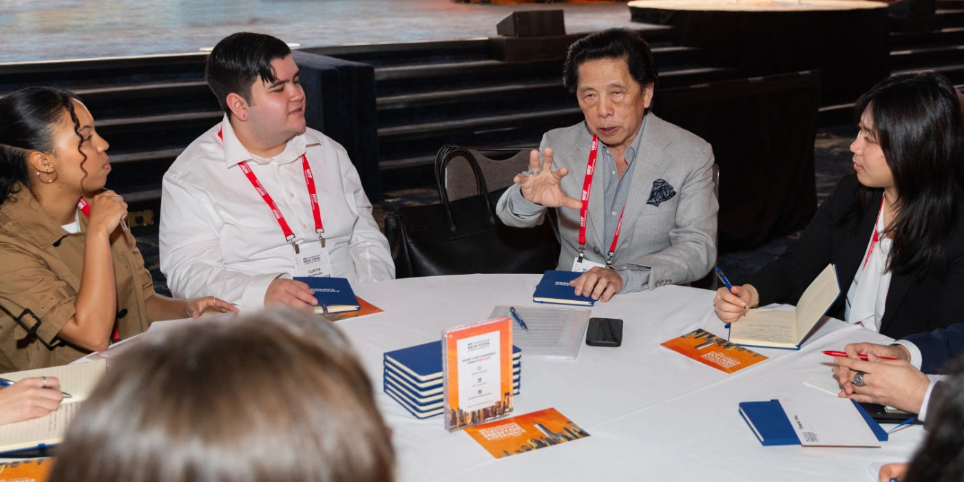 A group of people sit around a conference table, engaged in discussion with John Jay. They are taking notes in notebooks and conference materials on the table.