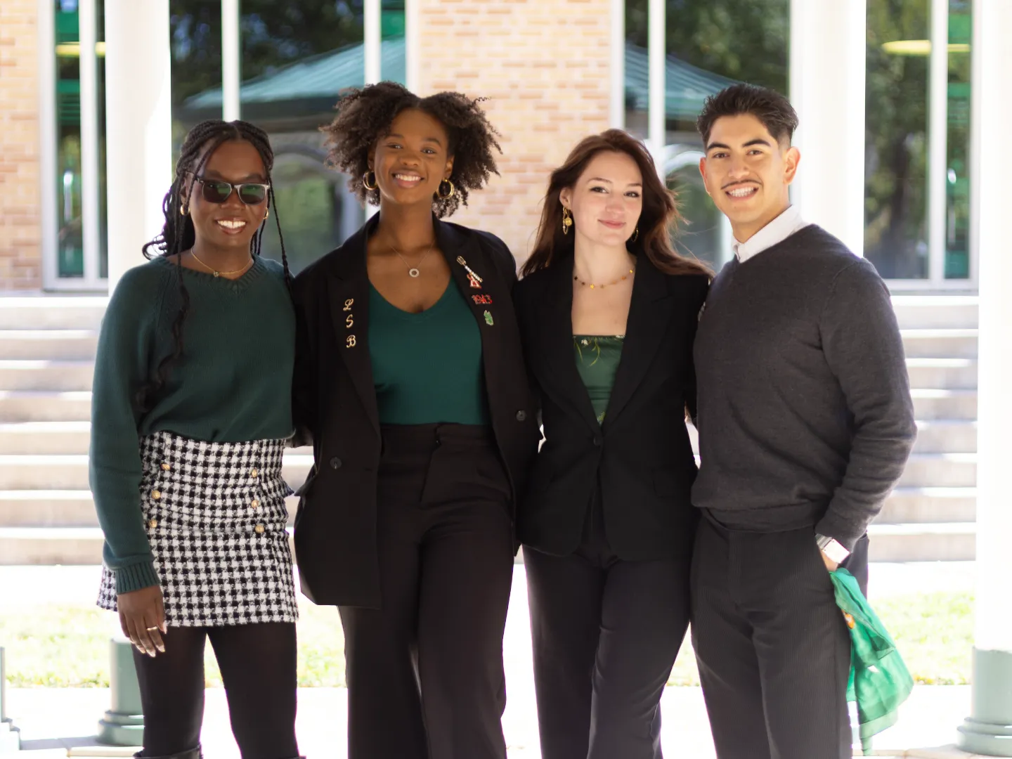 Four people standing together smiling, wearing semi-formal attire with a mix of green and black clothing, in front of a building with steps.