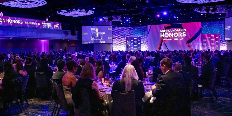A photograph of the NRF Foundation Honors ballroom. Hundreds of retail industry executives and managers are seated at round tables. A speaker is on stage with a purple and blue backdrop that reads NRF Foundation Honors.