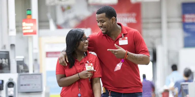 Two associates at a BJ's Wholesale Club take a moment to laugh with one another while walking the warehouse floor. The man on the right has his arm loosely around the women's shoulders while they smile and laugh.