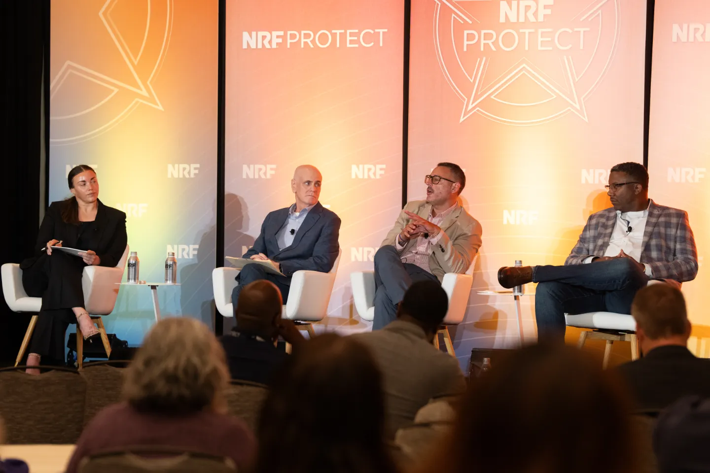 Panel discussion at NRF Protect conference with four speakers seated on stage in white chairs, speaking to an audience in front of large NRF Protect branded backdrops with warm orange lighting.
