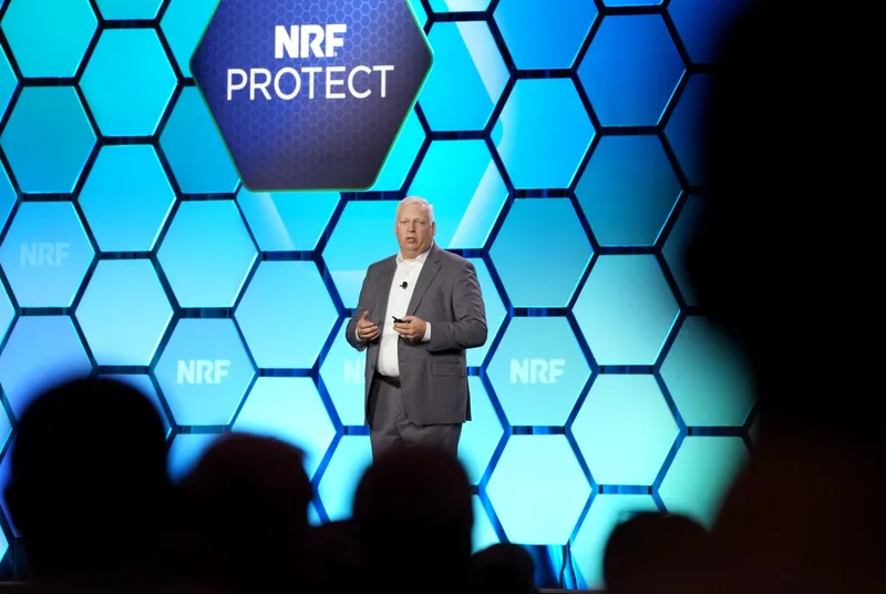Speaker presenting on stage at NRF PROTECT event with blue hexagonal backdrop and audience in foreground.