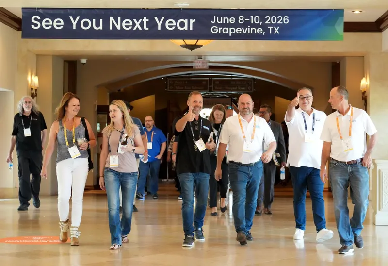 NRF PROTECT 2025 event sign with bold lettering on a dark background hangs above a hallway. A dozen men and women are walking towards the camera in the hallway. A man in a black polo-style shirt is giving a thumbs up gesture.