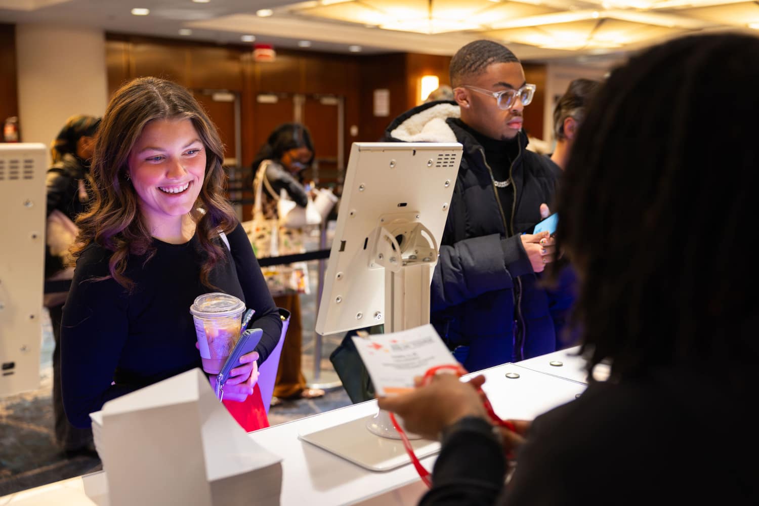 Two Student Program attendees check-in to the event, acquiring their badges representative standing next to an electronic kiosk.