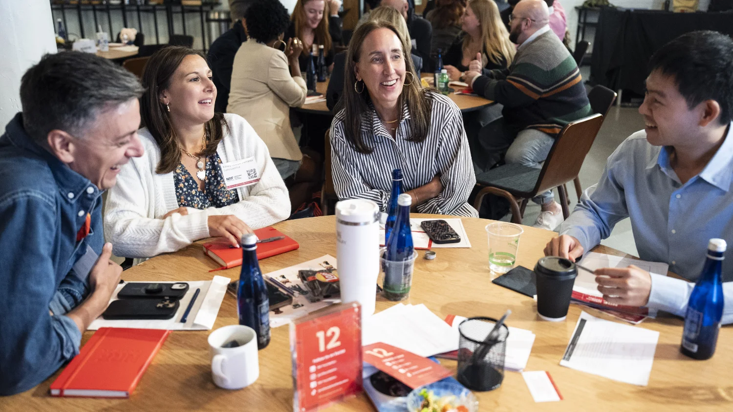 A group of people sit around a table laughing and engaging in conversation at a meeting or workshop, with drinks and notebooks scattered around.