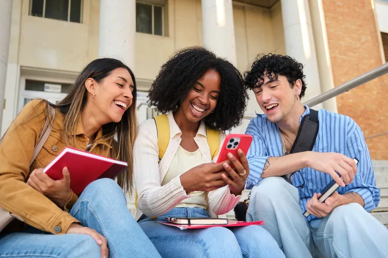 Three college students laugh while looking at a smartphone