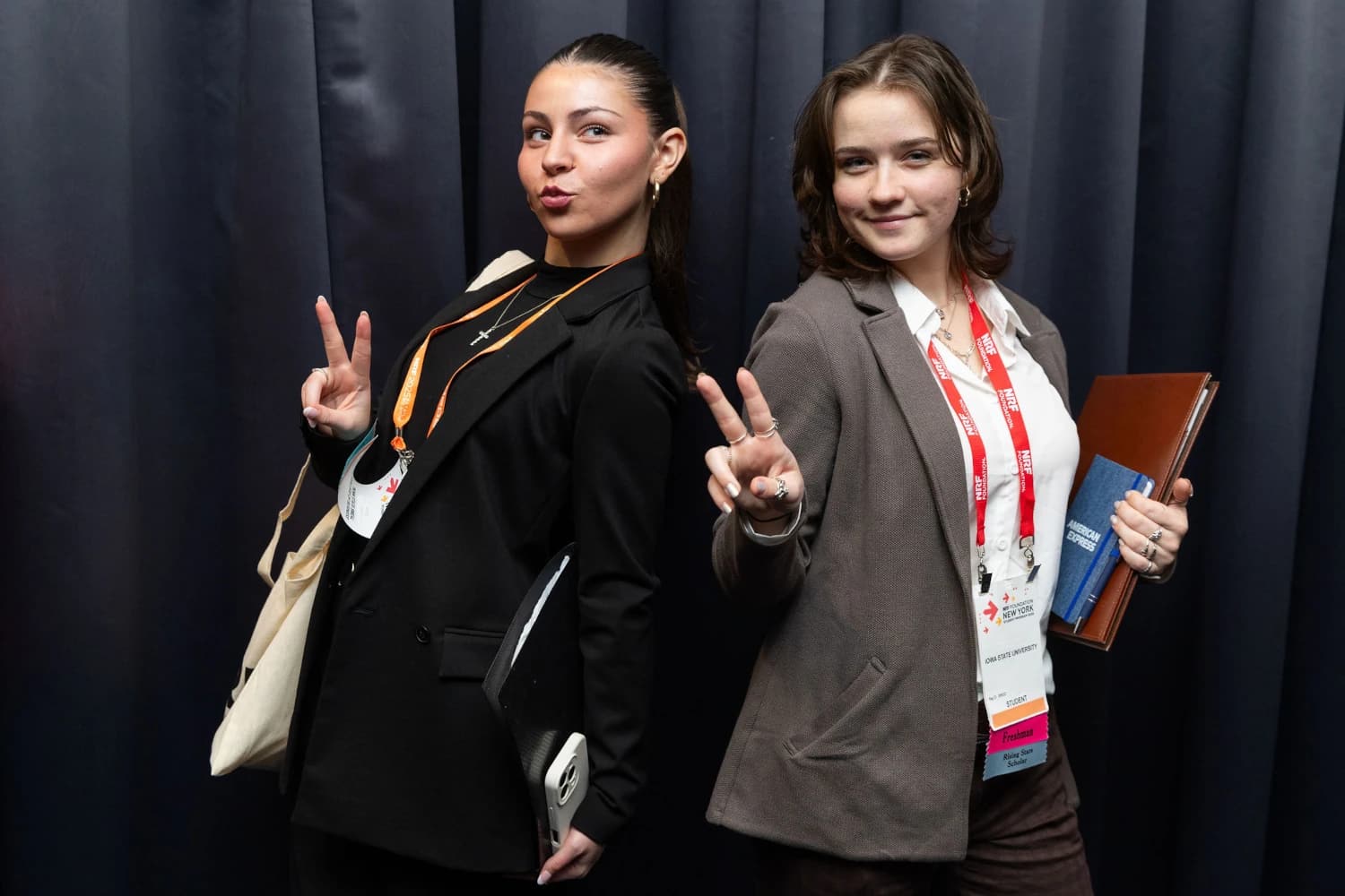 Two Student Program 2025 attendees pose for a photo during the career fair. They are holding materials for note taking and personal organization in one hand, while smiling and flashing a peace sign with the other hand.