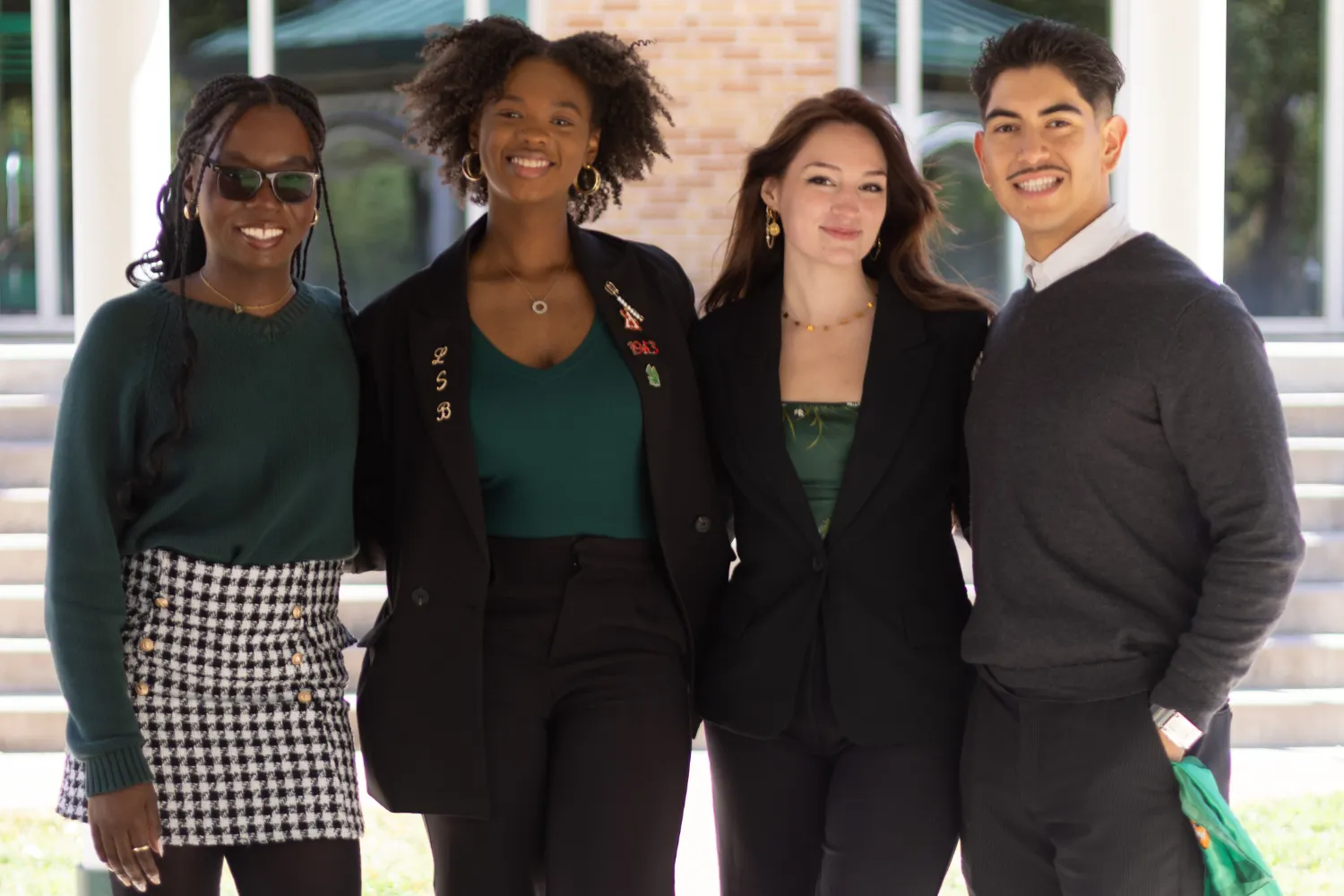 Four people smiling, standing together outdoors. They are wearing a mix of suits and casual attire with shades of green, and sunglasses.