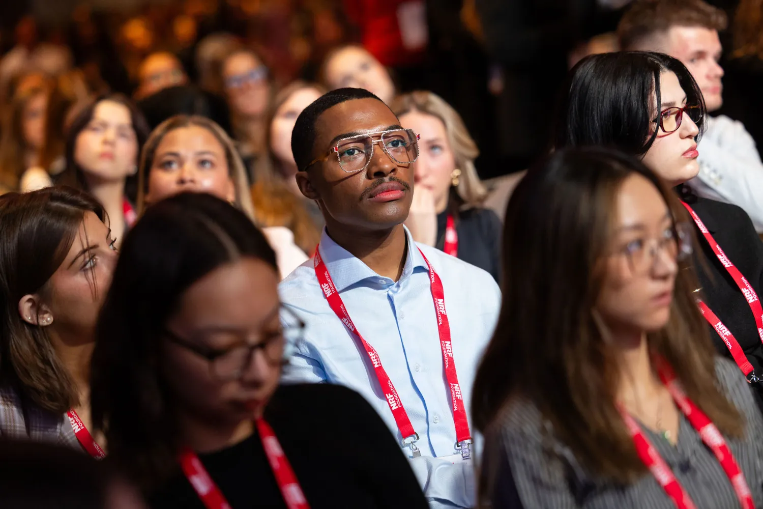 The Student Program 2025 audience attentively watches the speaker. In the center of the image is a young man wearing glasses rimmed with small gems.