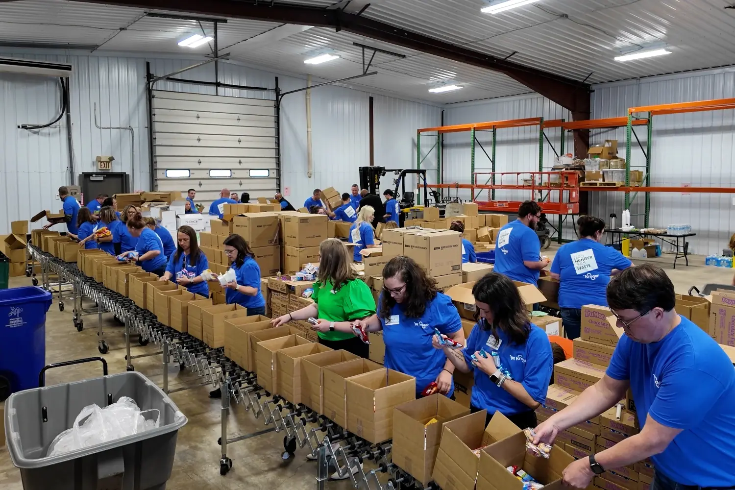 A group of Kroger associates in blue shirts pack food and supplies into cardboard boxes along a conveyor system inside a warehouse.